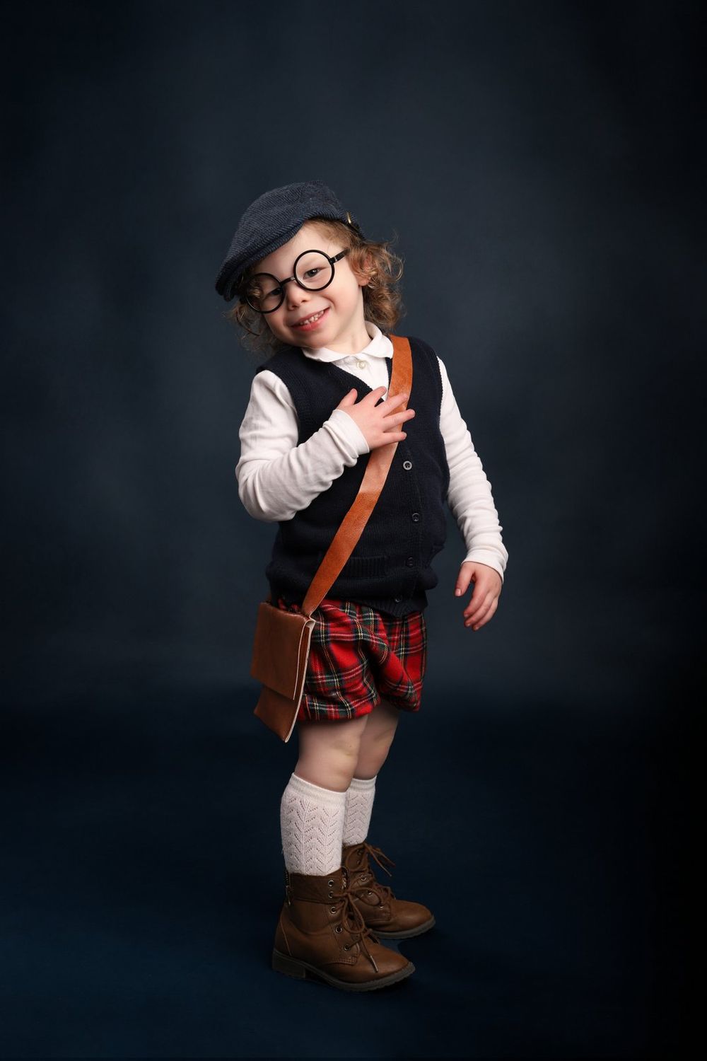 Small child dressed in vintage style with glasses, vest, plaid kilt, knee socks and leather boots poses on dark background.