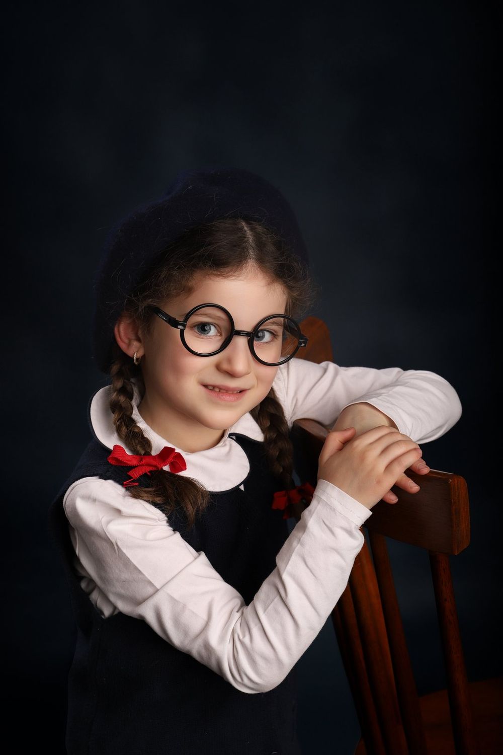 Student in round glasses and white shirt with red bows leans on wooden chair against dark background in portrait.