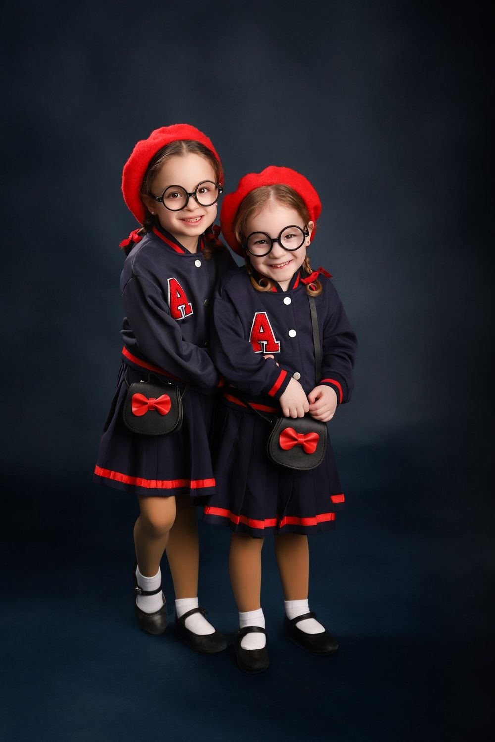 Two students wearing matching navy and red school uniforms with berets and glasses pose in front of dark backdrop.