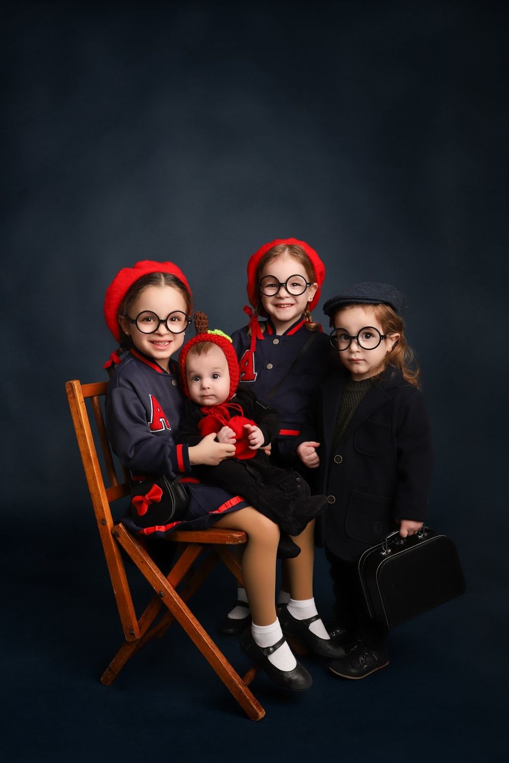 A group of children on a wooden chair wearing matching dark colored outfits with red accents and round glasses.
