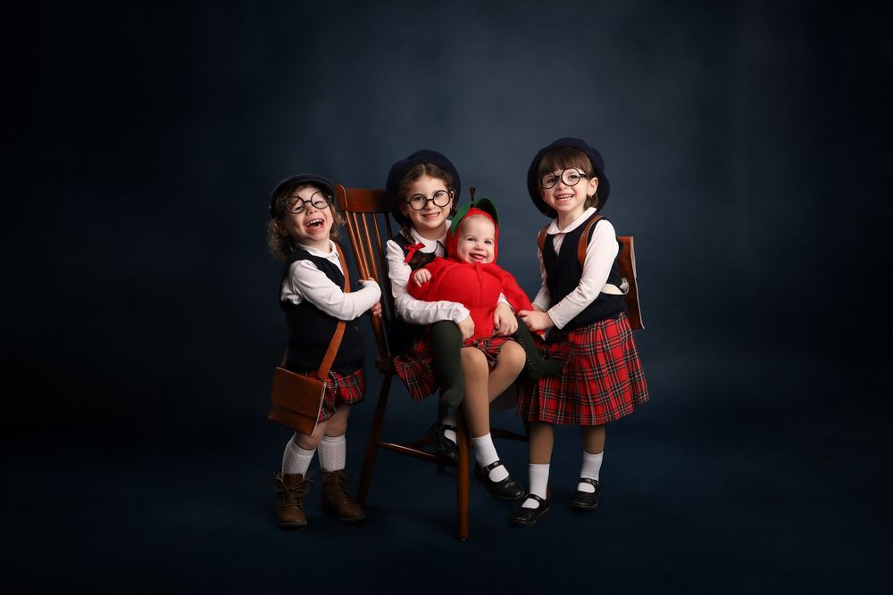 Three young children in traditional plaid school uniforms sit together on a wooden stool against a dark background.