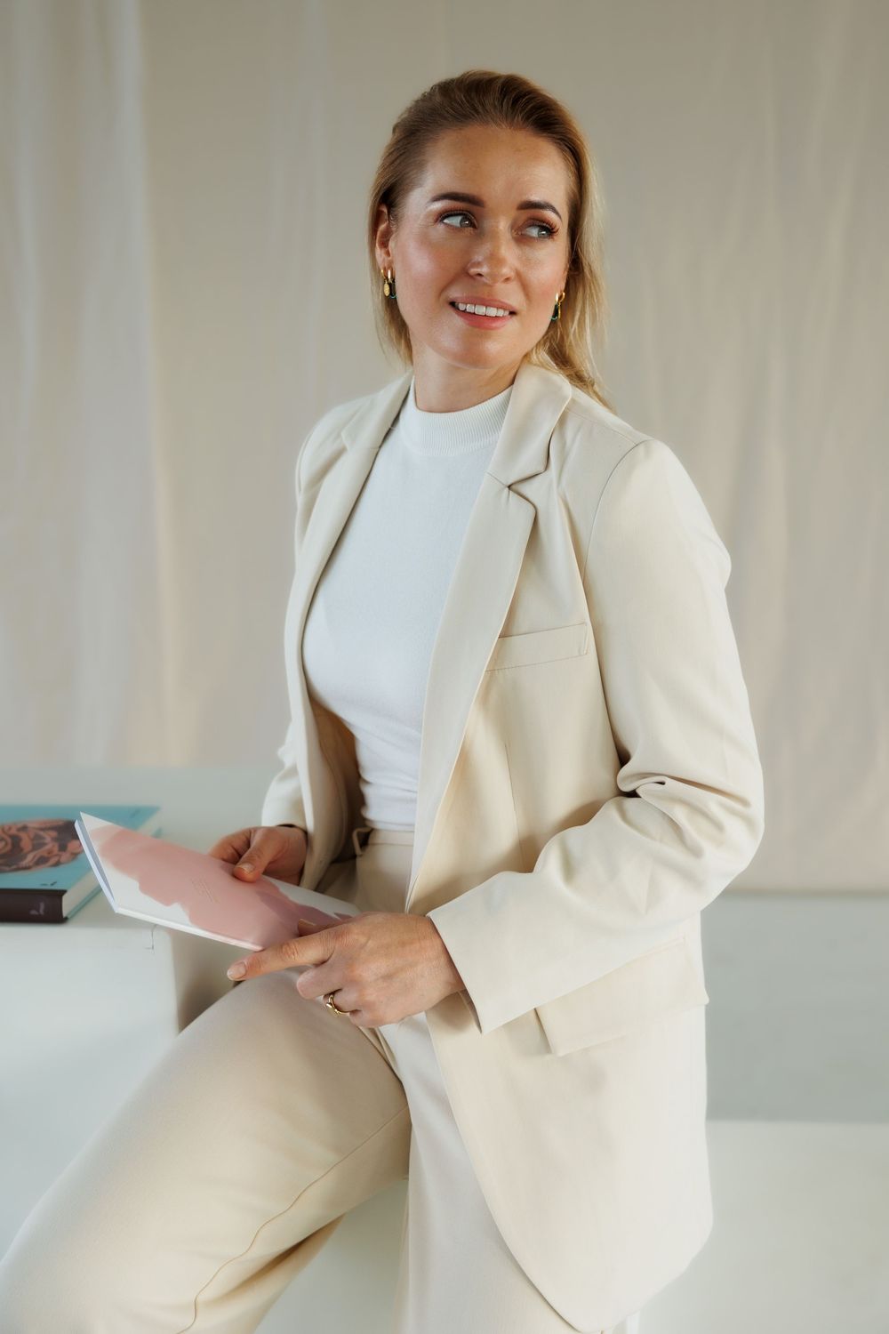 Professional woman in beige blazer holding tablet, smiling confidently in bright modern office setting.