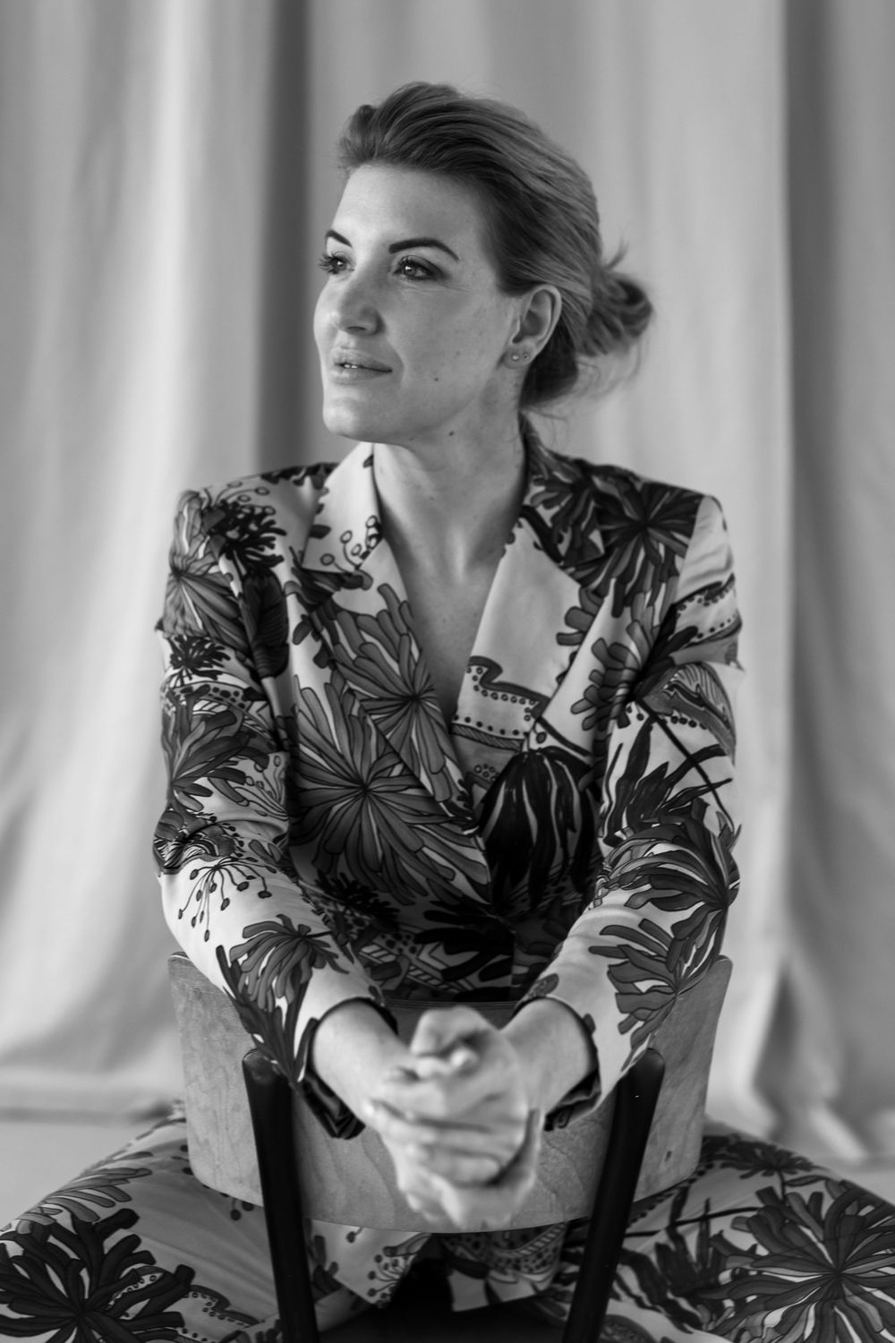 Black and white portrait of a woman in floral blazer seated, hands clasped, gazing thoughtfully to the side.