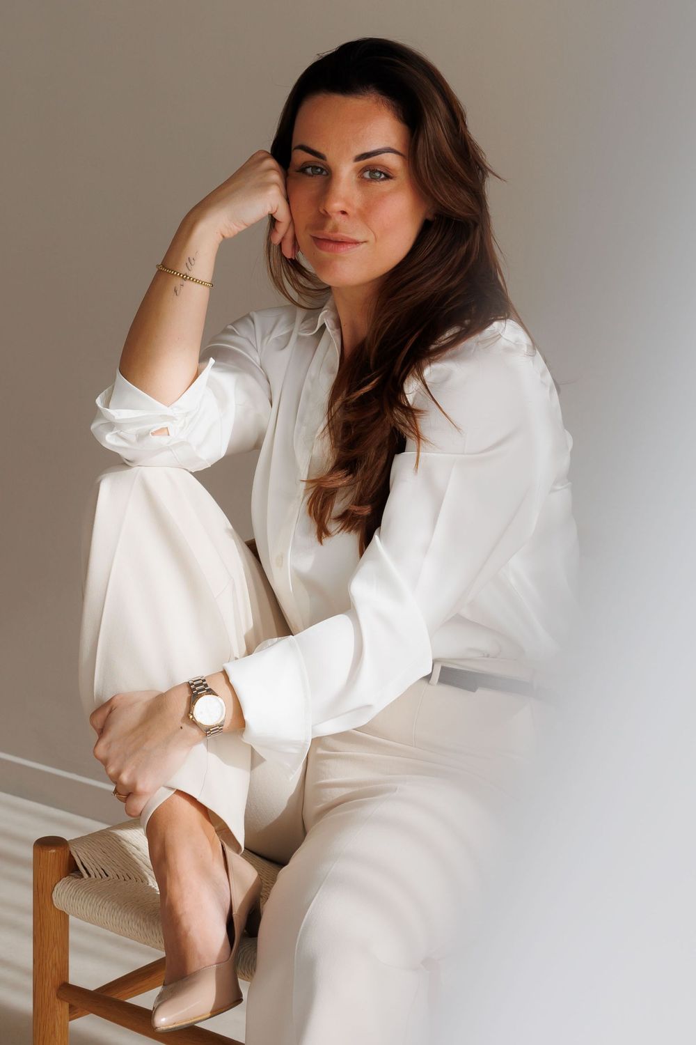 Woman in white blouse and trousers sits gracefully on wooden chair, resting head on hand, wearing gold watch.