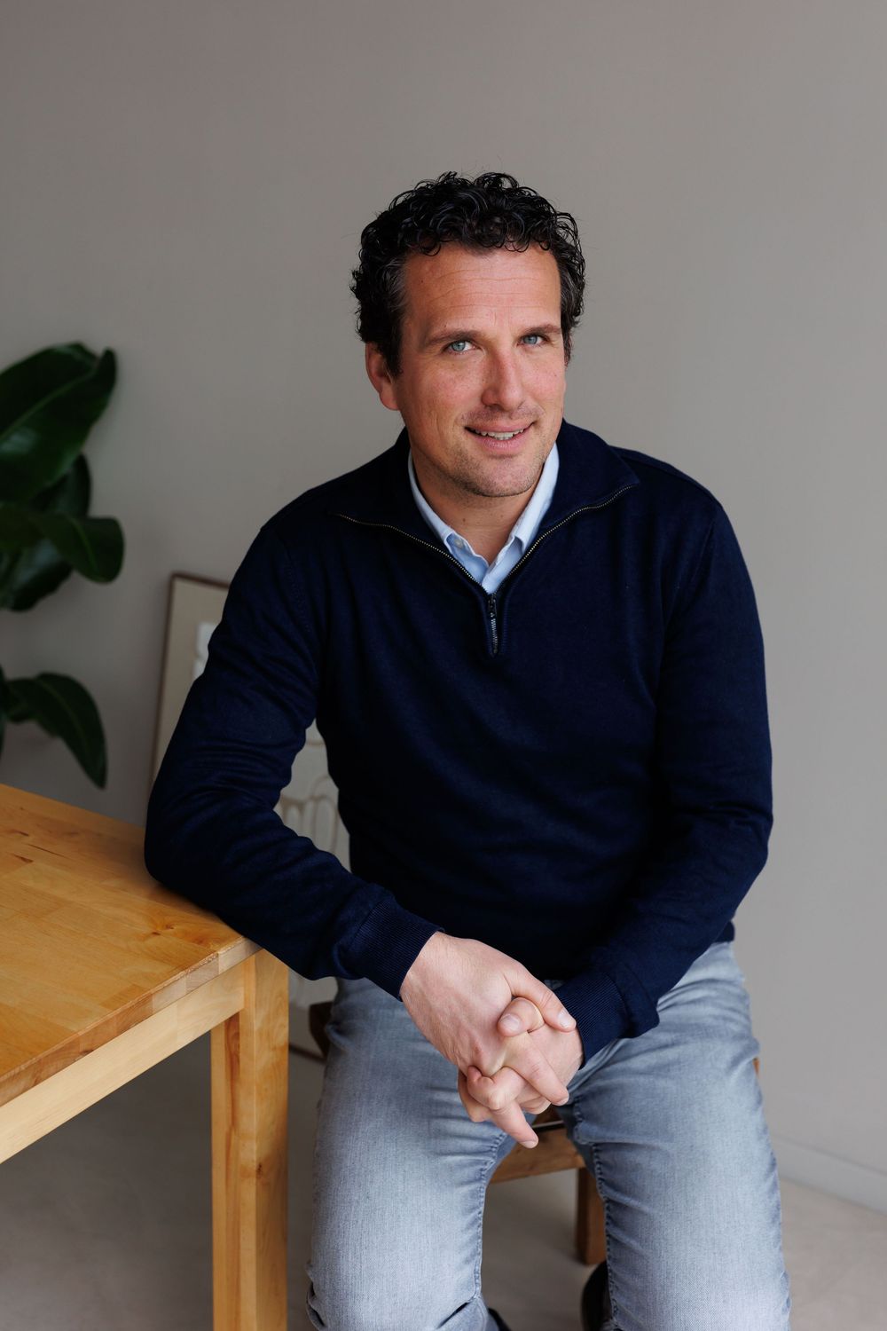 Man in navy sweater sits relaxed at wooden table, smiling confidently in a bright, modern minimalist space.