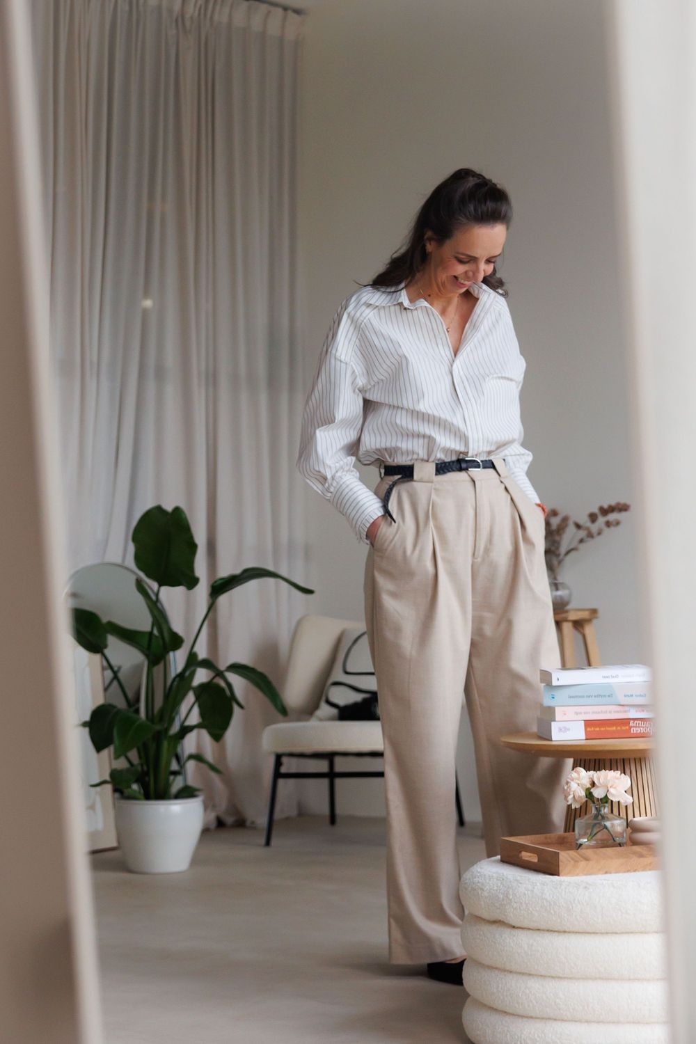 Woman in striped shirt and wide-leg beige trousers standing in a modern, minimalist living room with plants and books.