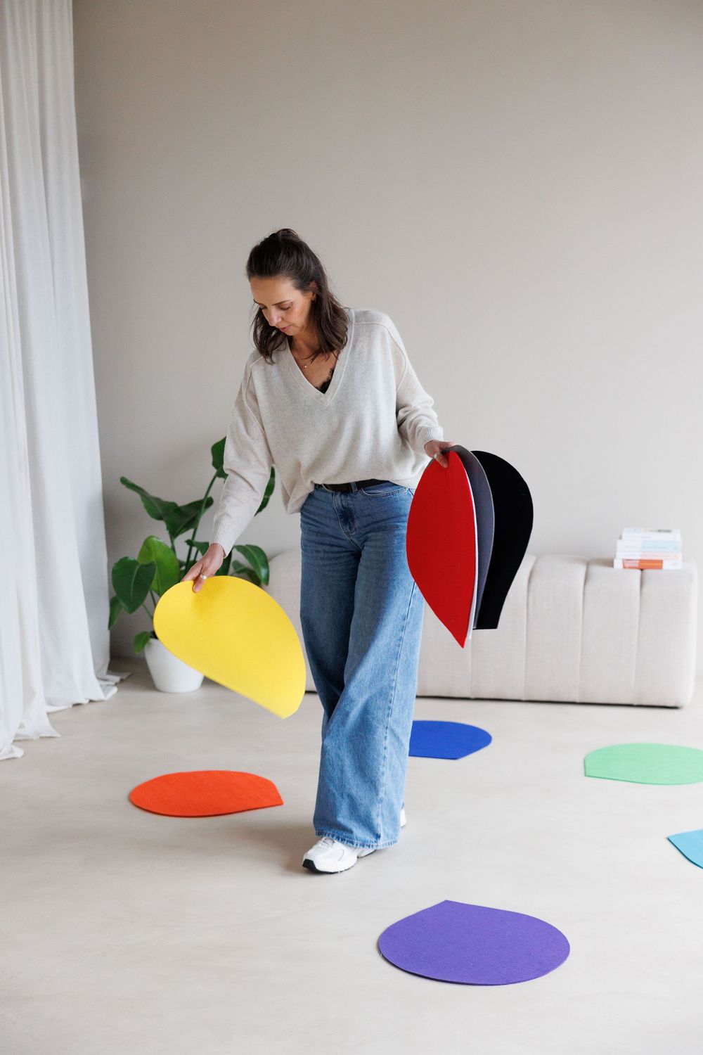 Woman arranging colorful felt shapes on a light wooden floor in a bright modern room.