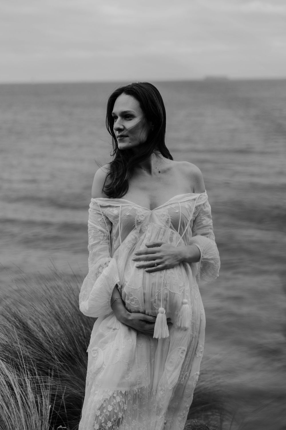 Elegant pregnant woman in sheer white off-shoulder dress cradles bump by ocean in black and white.