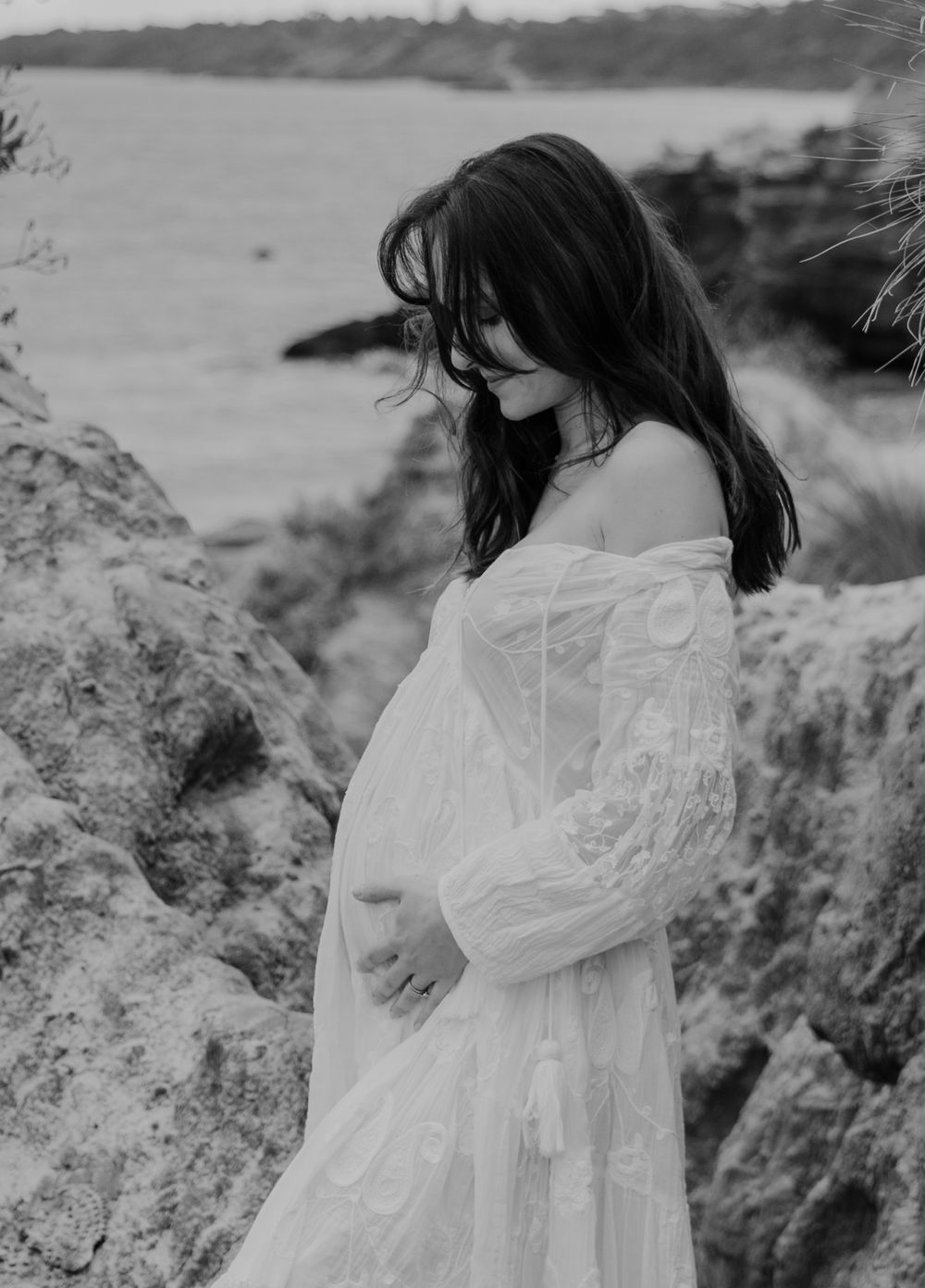 Pregnant woman in white lace off-shoulder dress holds bump near coastal rocks in black and white.