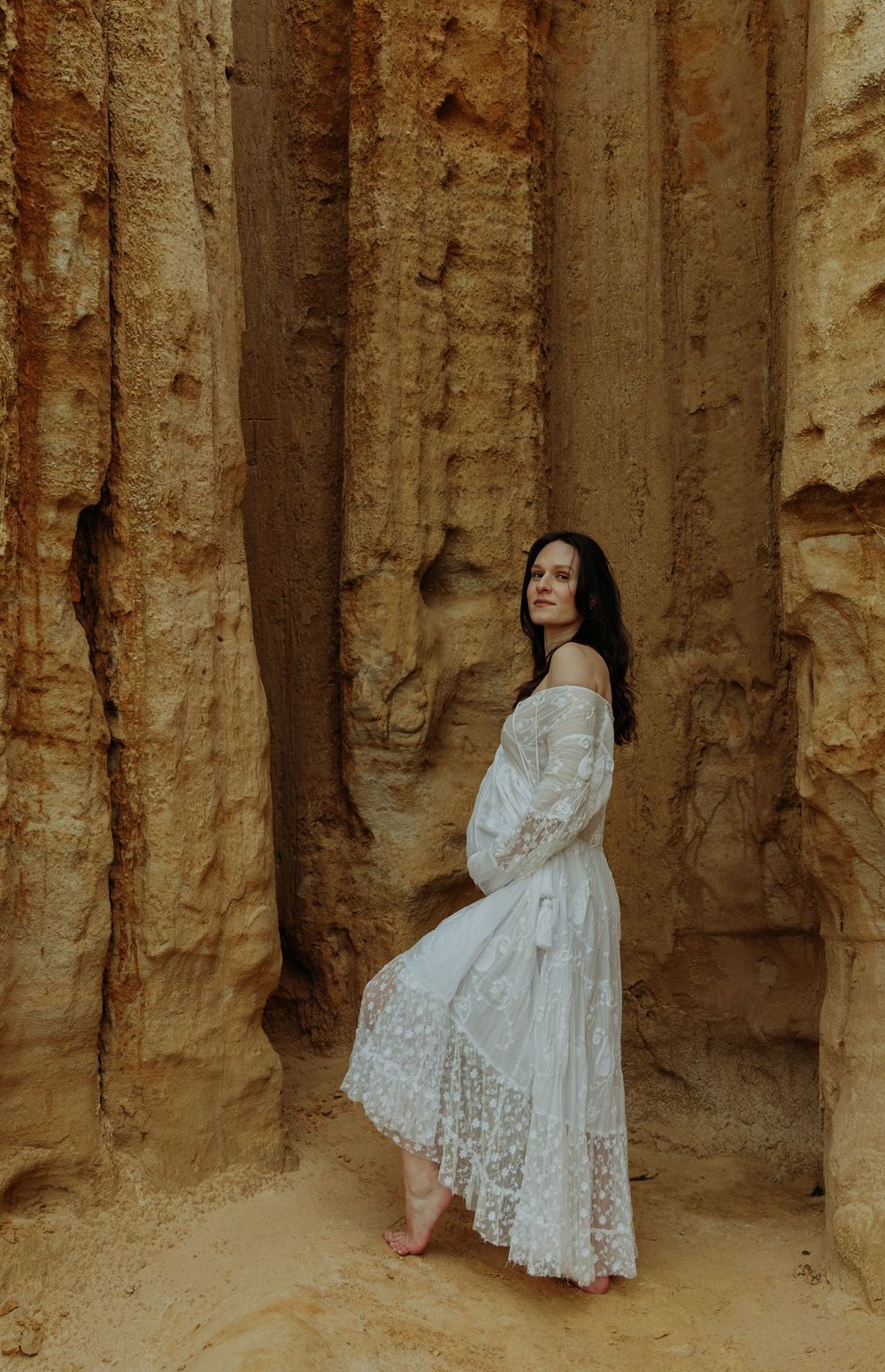 Pregnant woman in white lace off-shoulder dress standing between tall sandstone rock formations.
