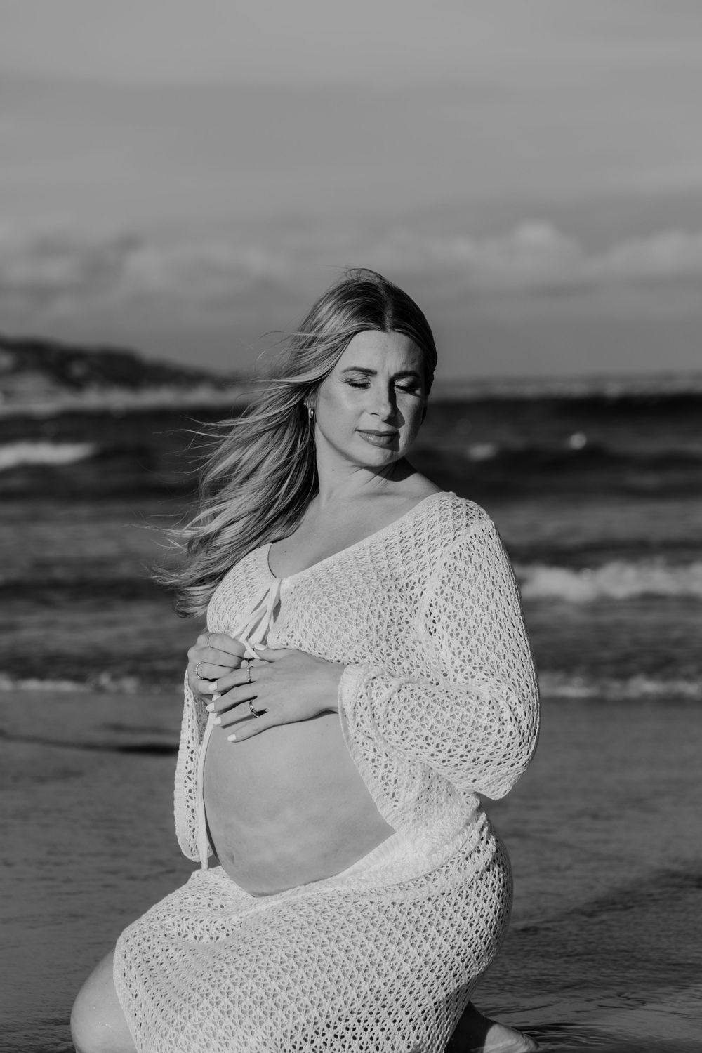 Black and white photo of pregnant woman in white crochet dress holding baby bump on beach.