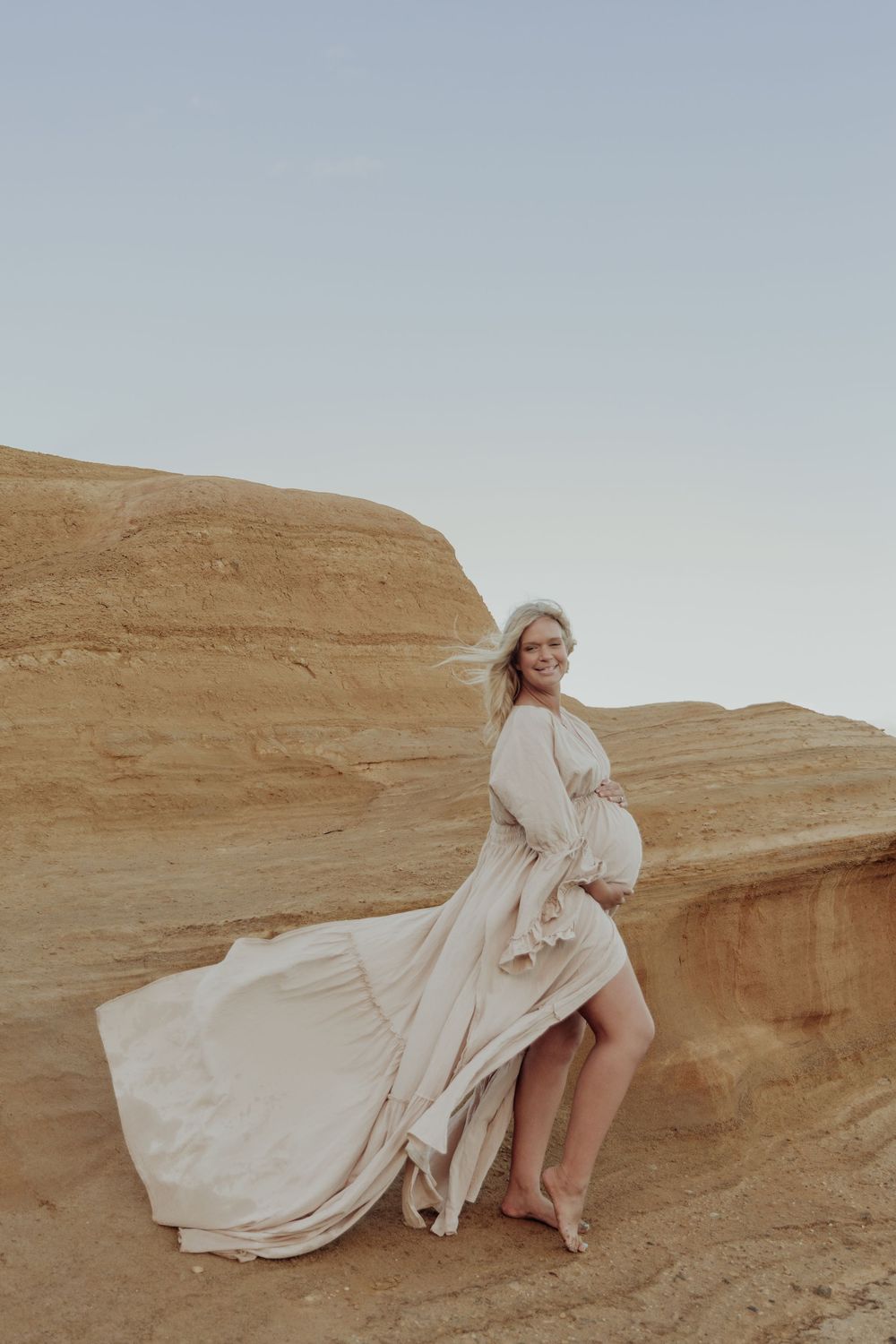 Pregnant woman in flowing cream dress smiling on sandy cliffs with dress billowing in the wind.