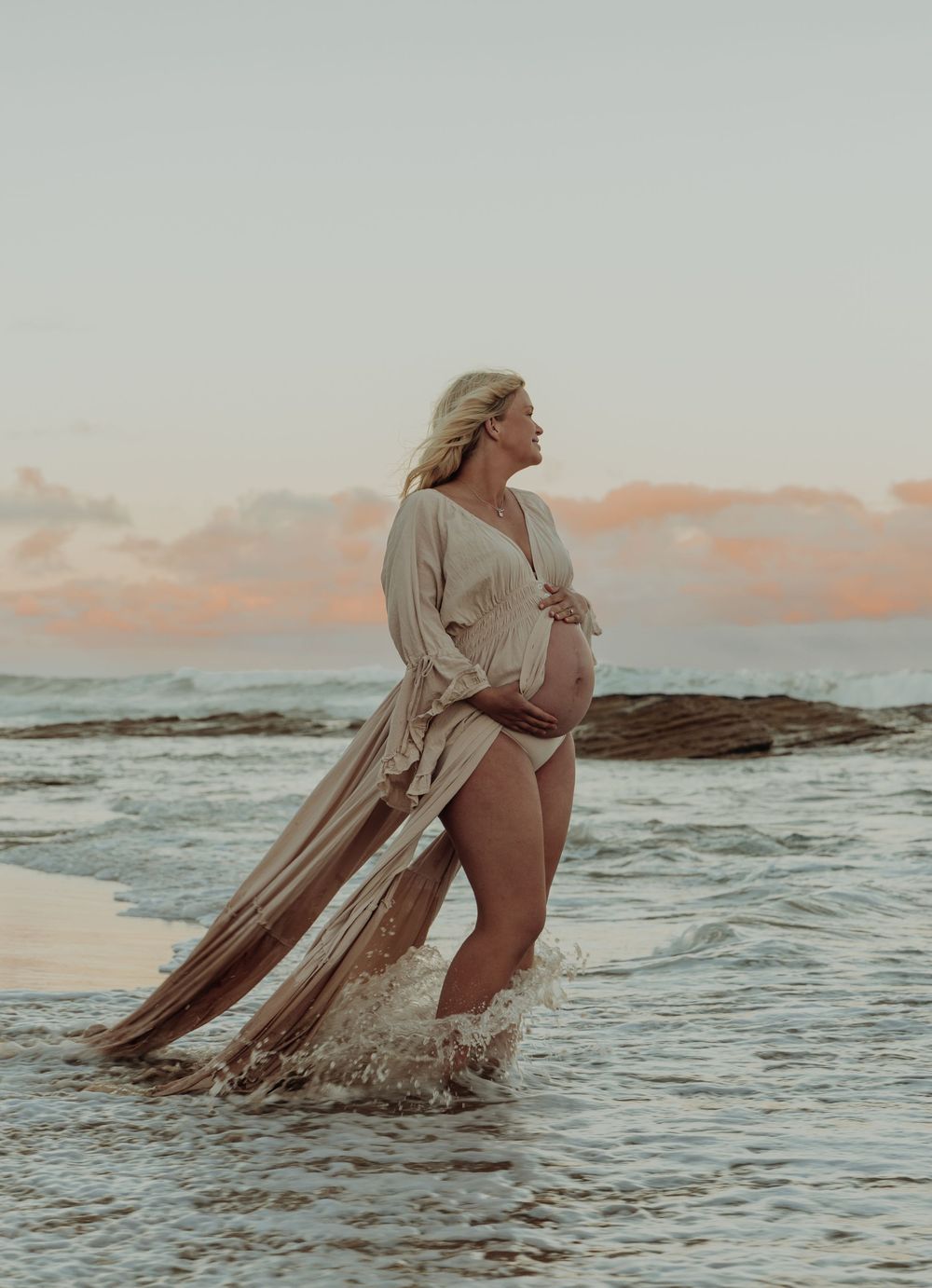 Pregnant woman in flowing open dress cradles bump while standing in ocean waves at sunset.