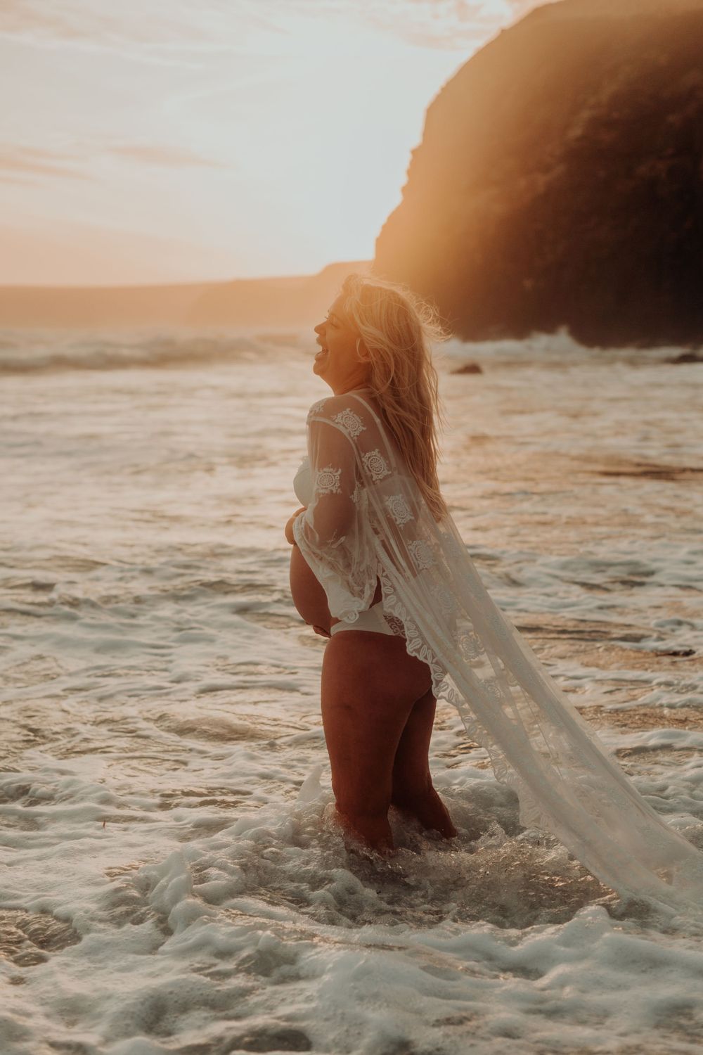 Pregnant woman in white lace robe standing in ocean waves at golden sunset.