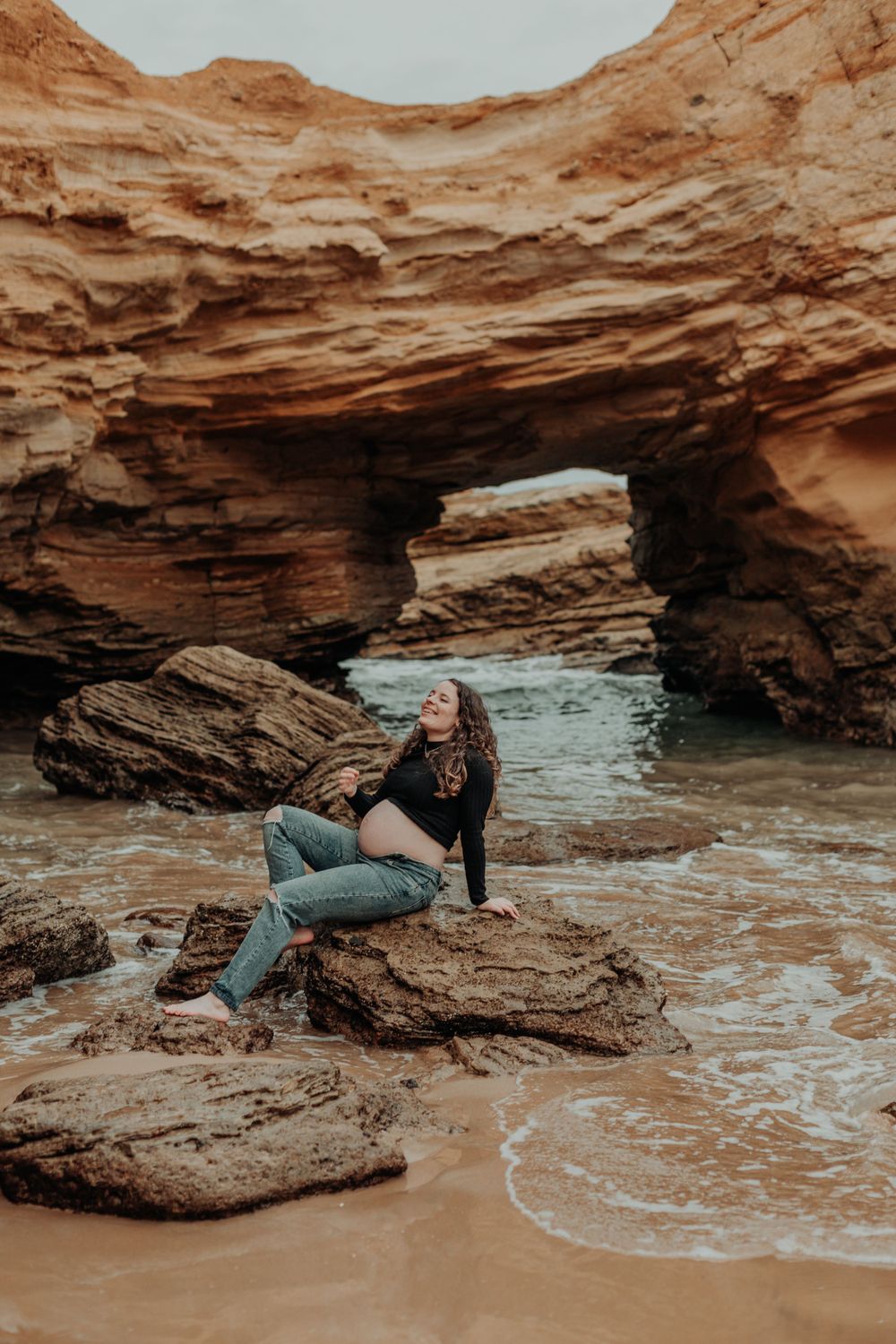 Pregnant woman in black top sitting on rocks near sandstone arch at beach.