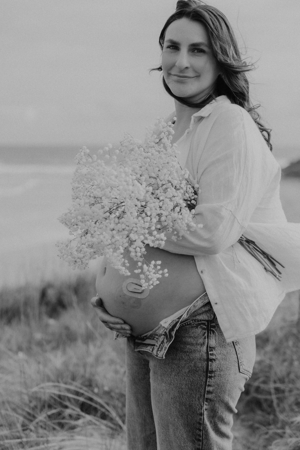 Black and white portrait of pregnant woman holding baby\u2019s breath flowers on coastal dunes.