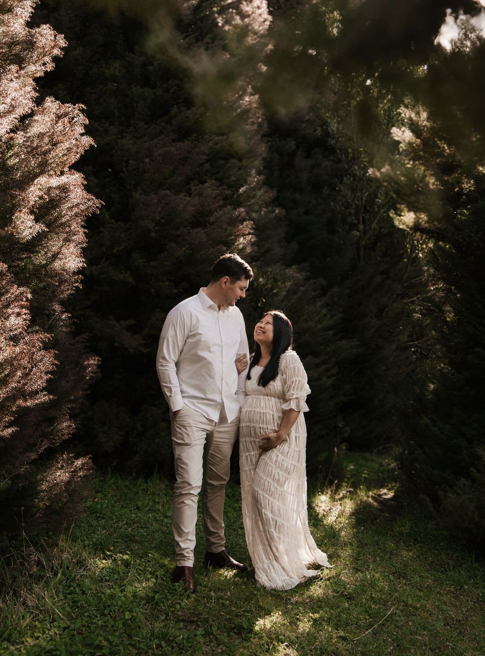 A couple in white attire stands among tall dark trees, the woman cradling her baby bump in a serene maternity photo.