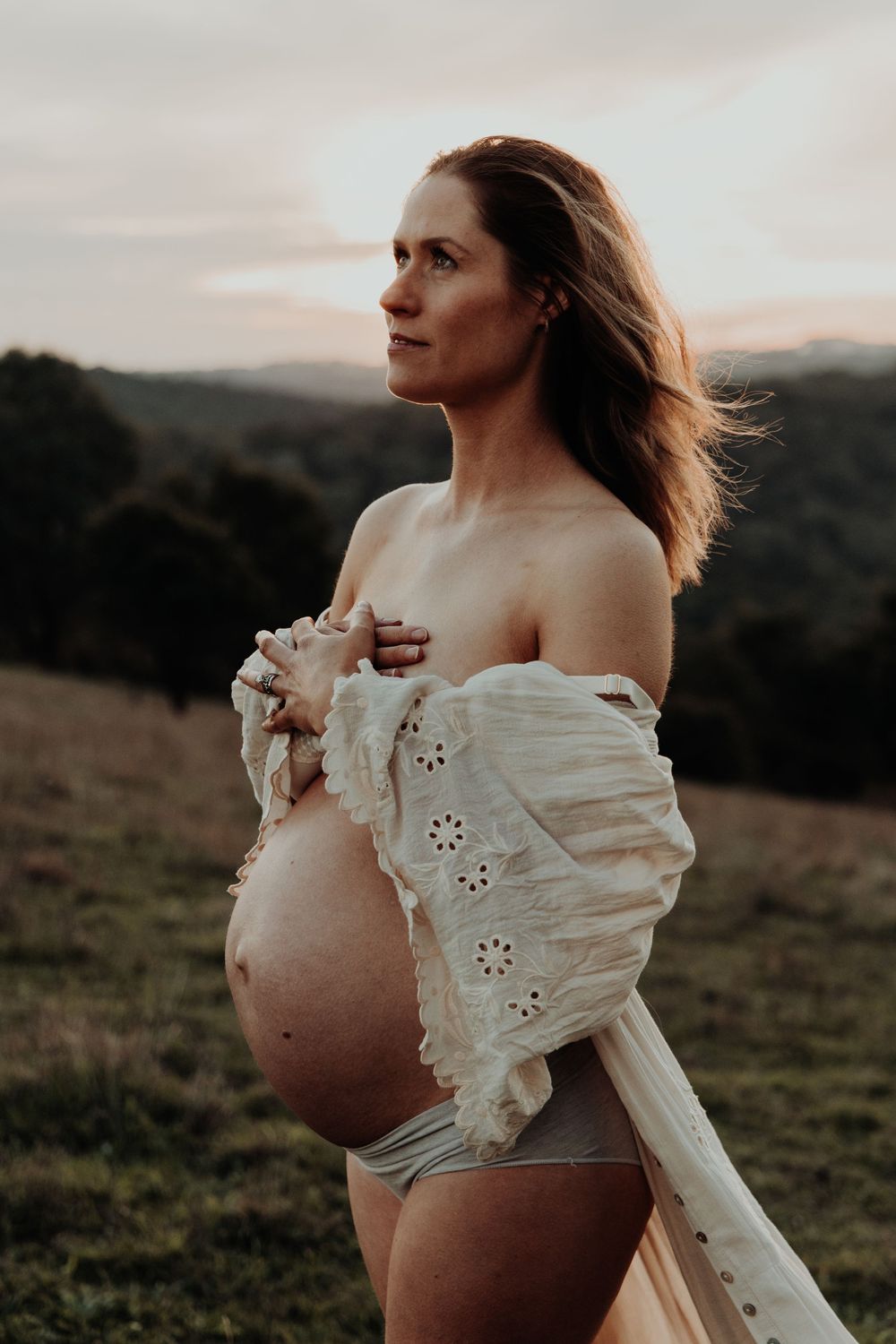 Pregnant woman holds white lace dress off shoulders revealing bare bump at golden hour on hillside.