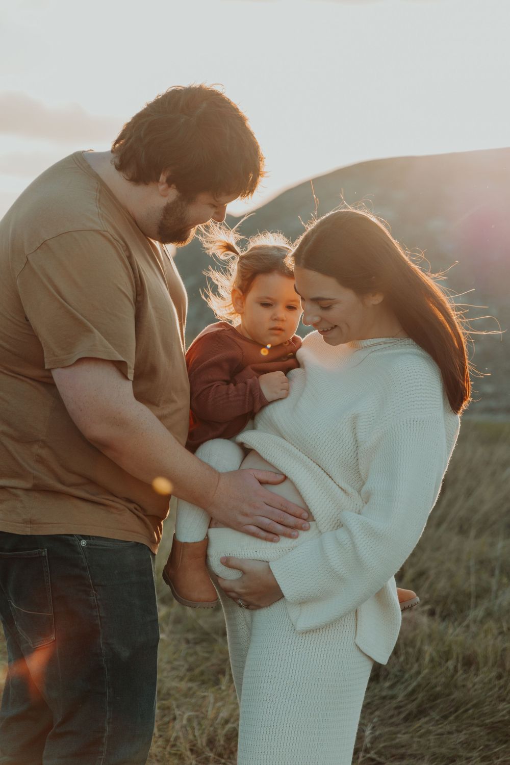 Family maternity photo at sunset, man holding toddler while pregnant woman cradles bump.