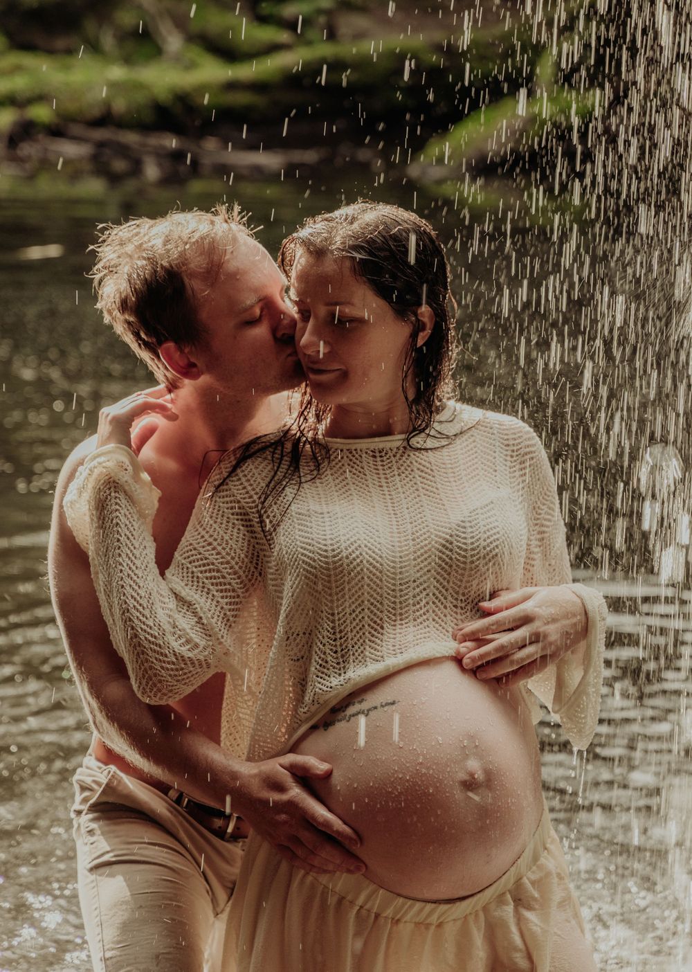 Couple embracing in rain during maternity shoot, man kissing pregnant woman's cheek outdoors.