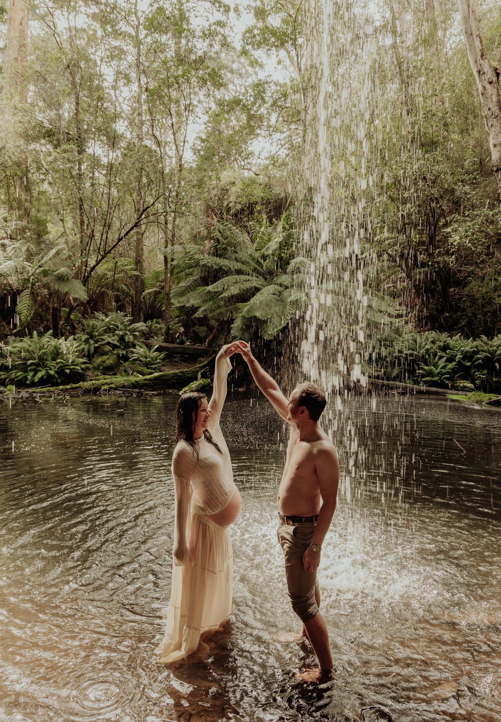 Couple dancing under waterfall in lush forest, pregnant woman in white dress, maternity session.