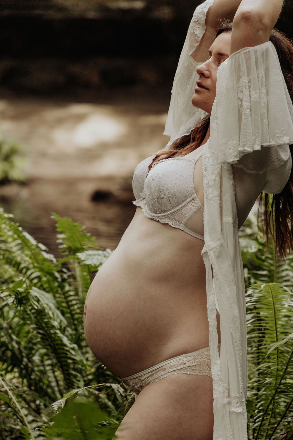 Pregnant woman in white lace lingerie and robe posing among lush green ferns near waterfall.