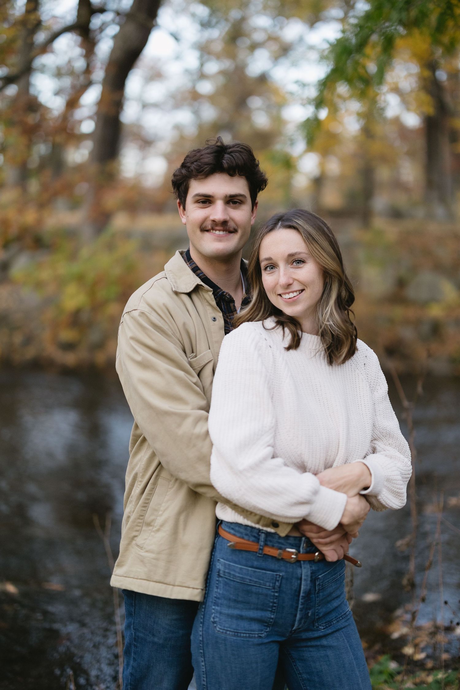 A couple posing together near a lake with autumn foliage in the background.