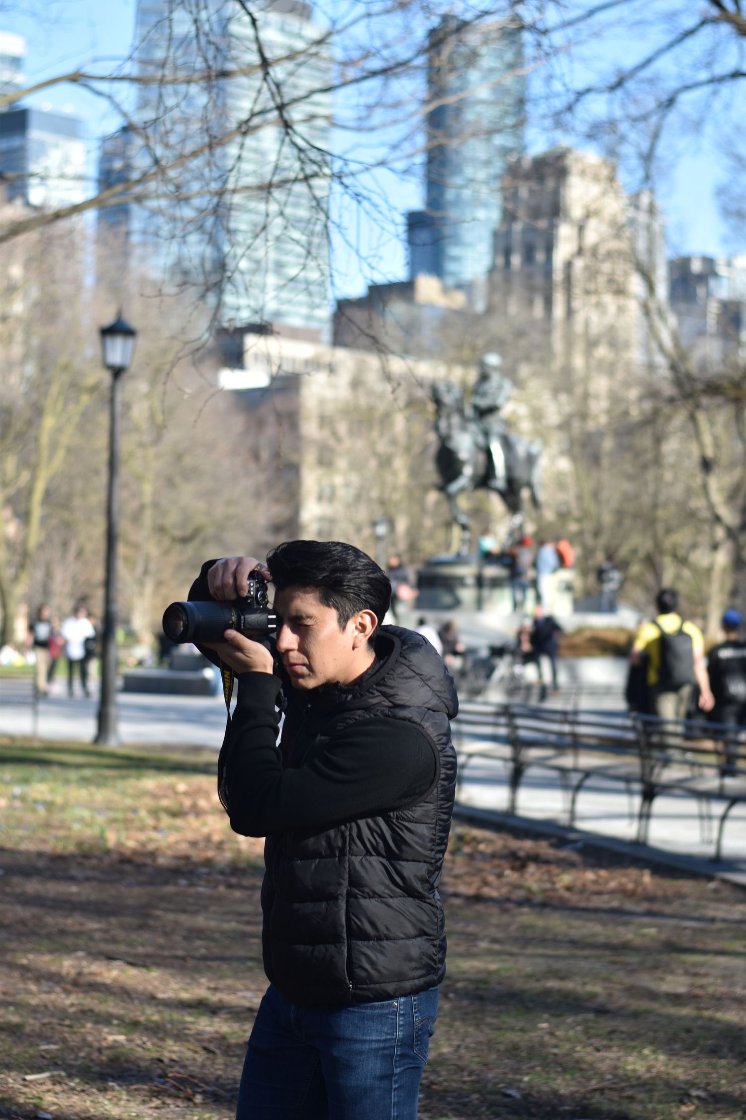 Man in black puffer jacket photographs with DSLR camera in a sunny urban park setting. Toronto GTA