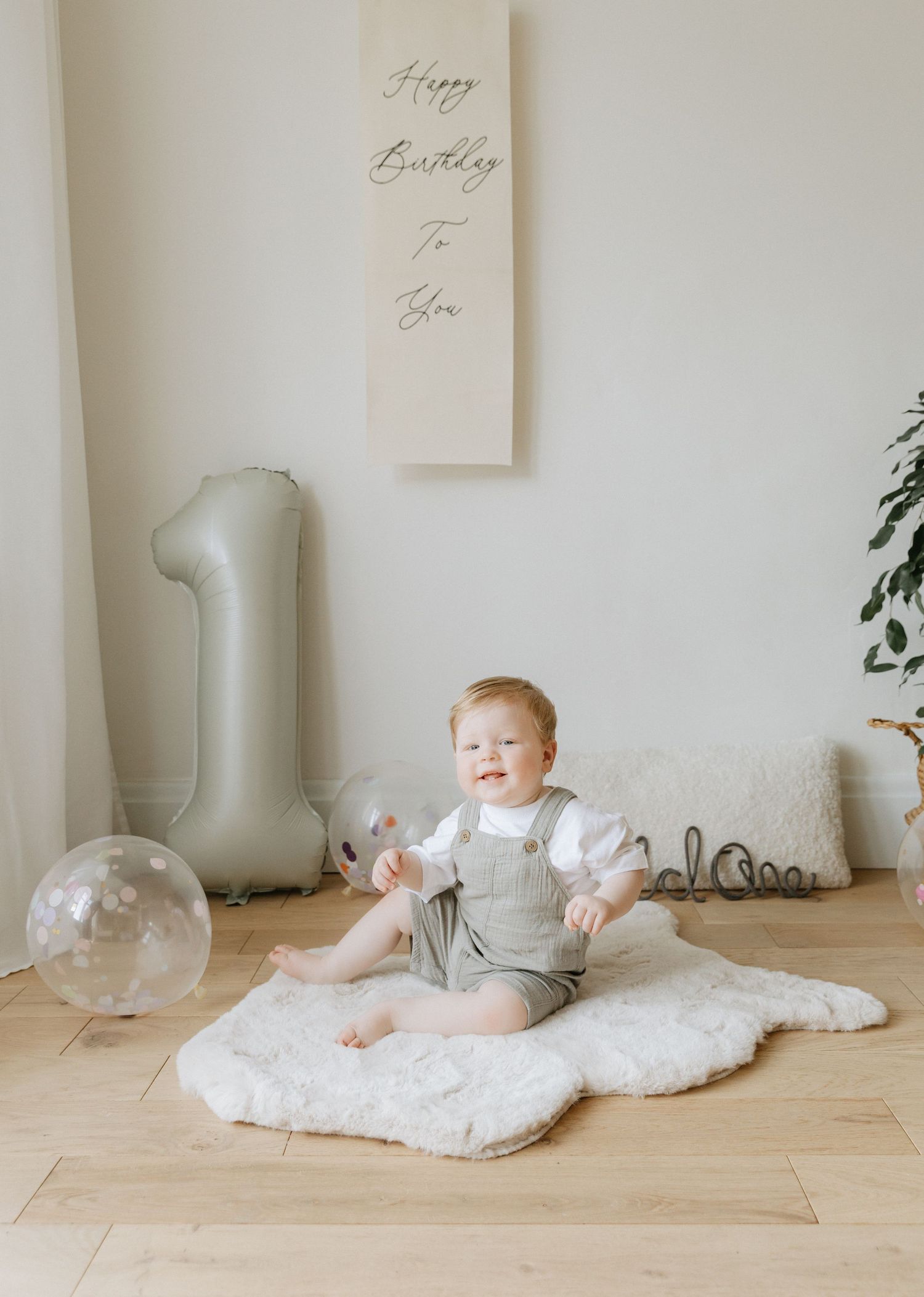 Baby sitting on white blanket in minimalist room with decorative balloons.