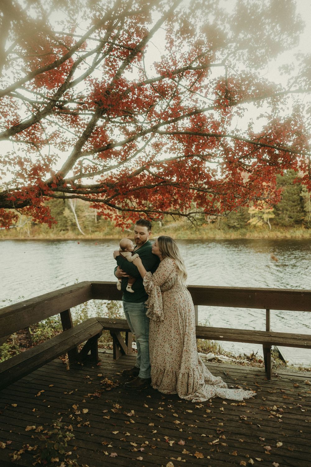 A couple stands with their baby on a wooden dock under vibrant red fall foliage.