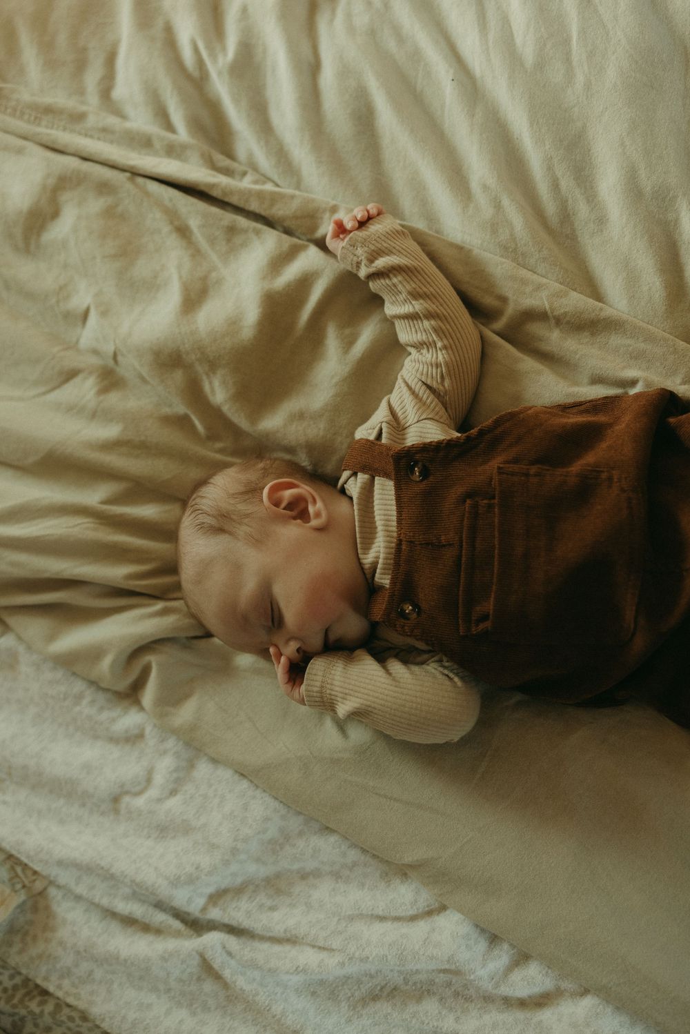 A small child rests peacefully on cream-colored bedding wearing brown clothing.