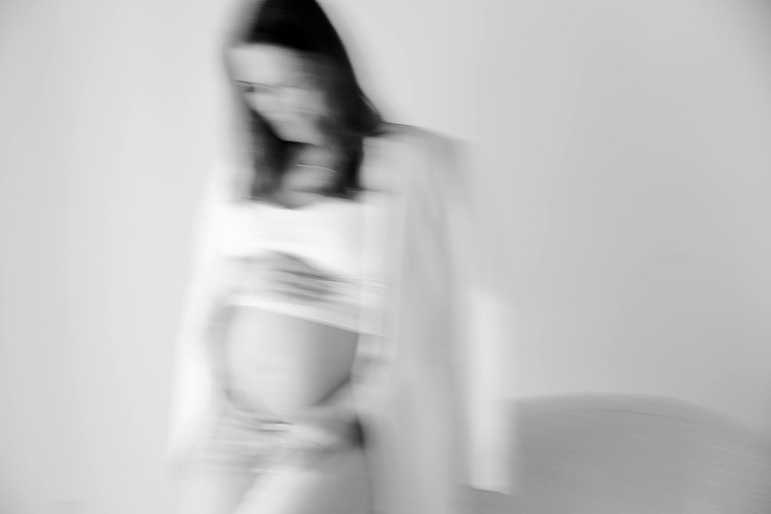 Gravid kvinde i hvid blazer og jeans holder om maven, stående ved vinduet i et lyst, minimalistisk rum. Fotograf i Middelfart.