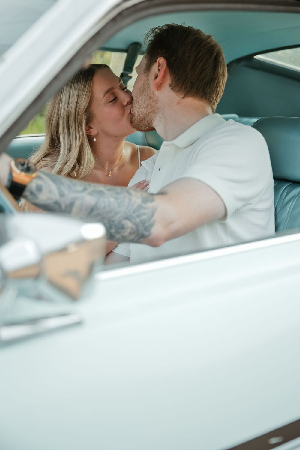 A couple shares a romantic kiss inside a vintage car, the man's tattooed arm visible on the steering wheel.
