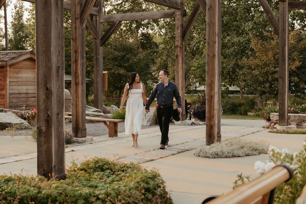 Couple walks hand-in-hand under rustic wooden pergola in a garden setting, woman in white dress, man in dark attire.