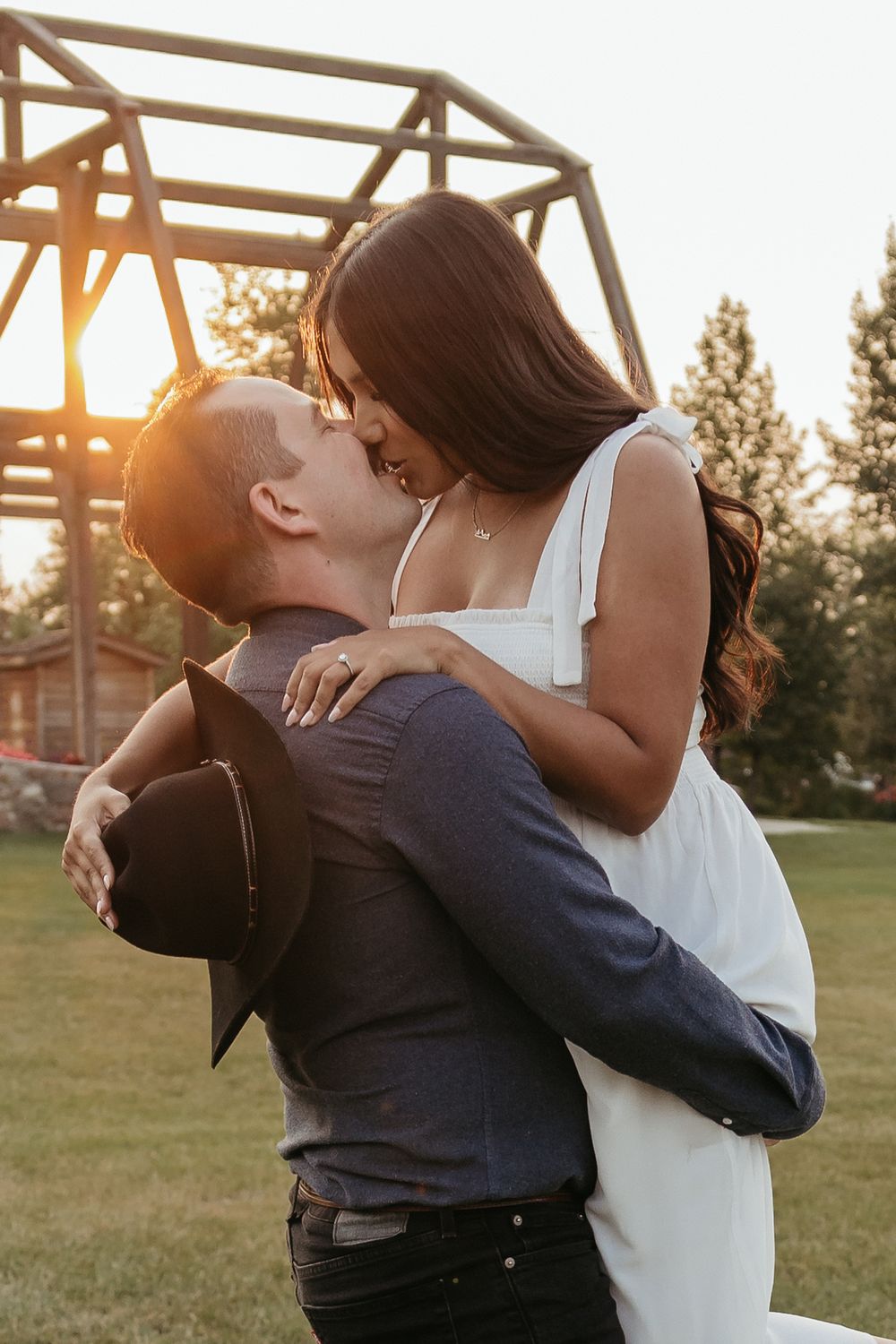 Couple sharing a romantic kiss at sunset, man lifting woman in white dress outdoors near wooden structure.