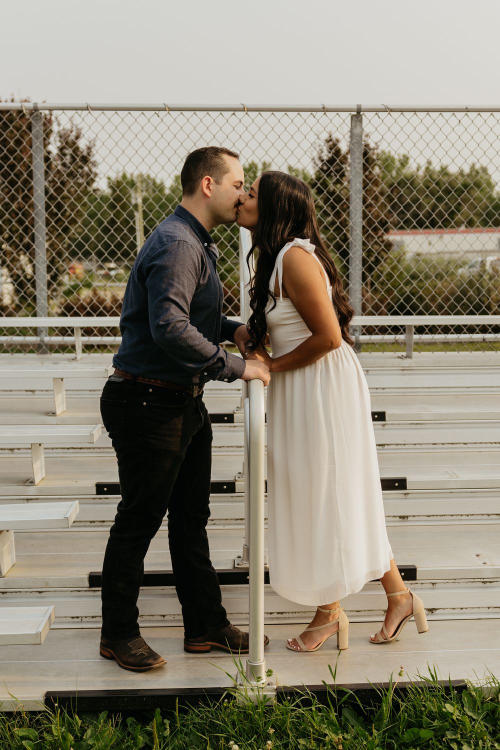 Couple sharing a romantic kiss on bleachers, man in blue shirt and woman in white dress, outdoor setting.