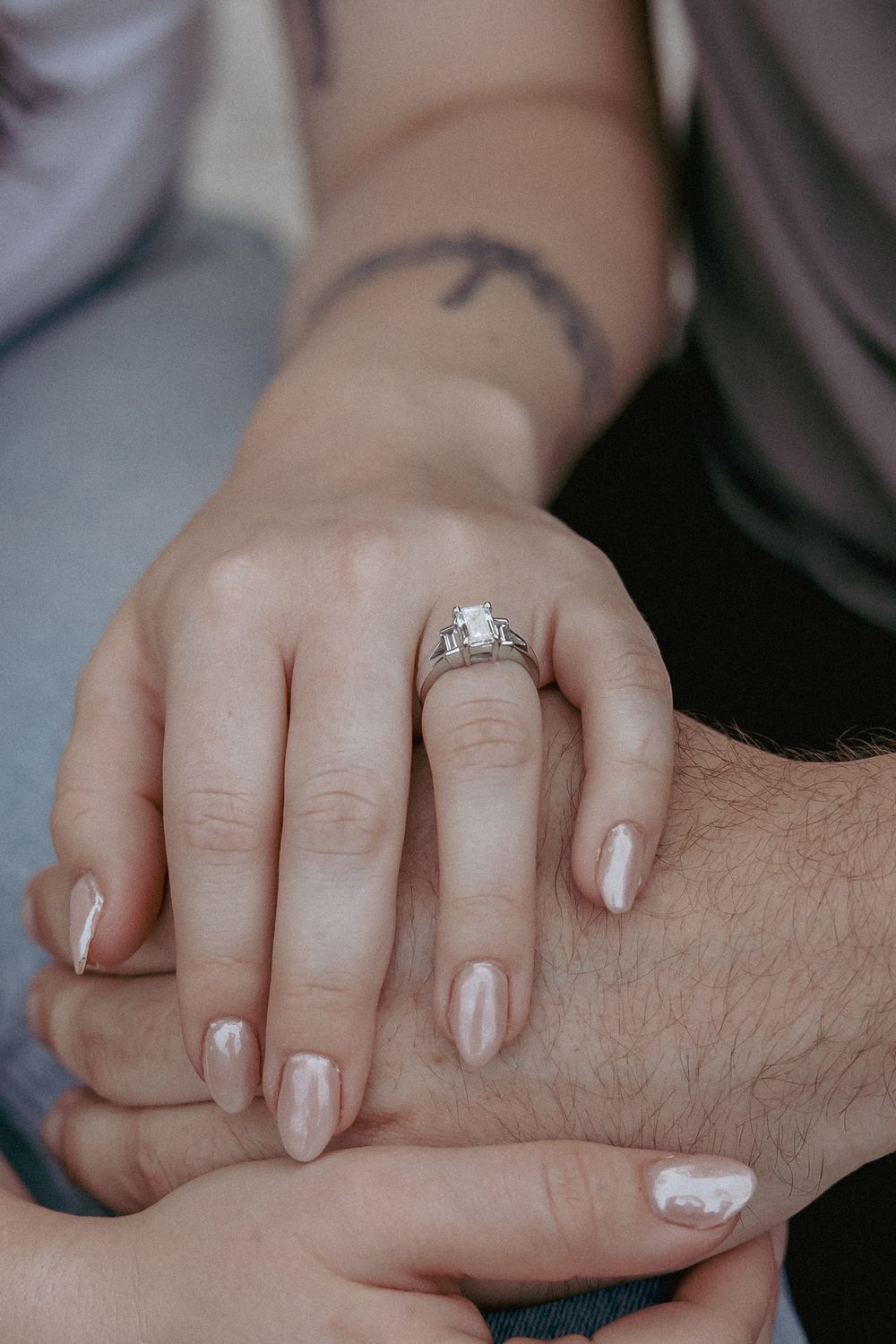 Close-up of a woman's hand with a diamond engagement ring and nude nails resting on a man's hand.