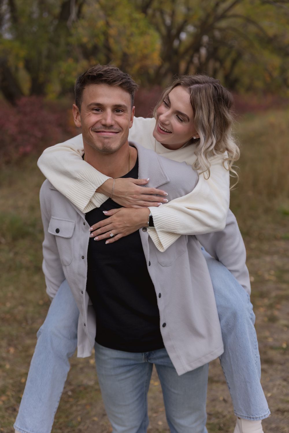 Smiling couple in autumn park, woman hugging man from behind, both wearing white jackets and jeans.