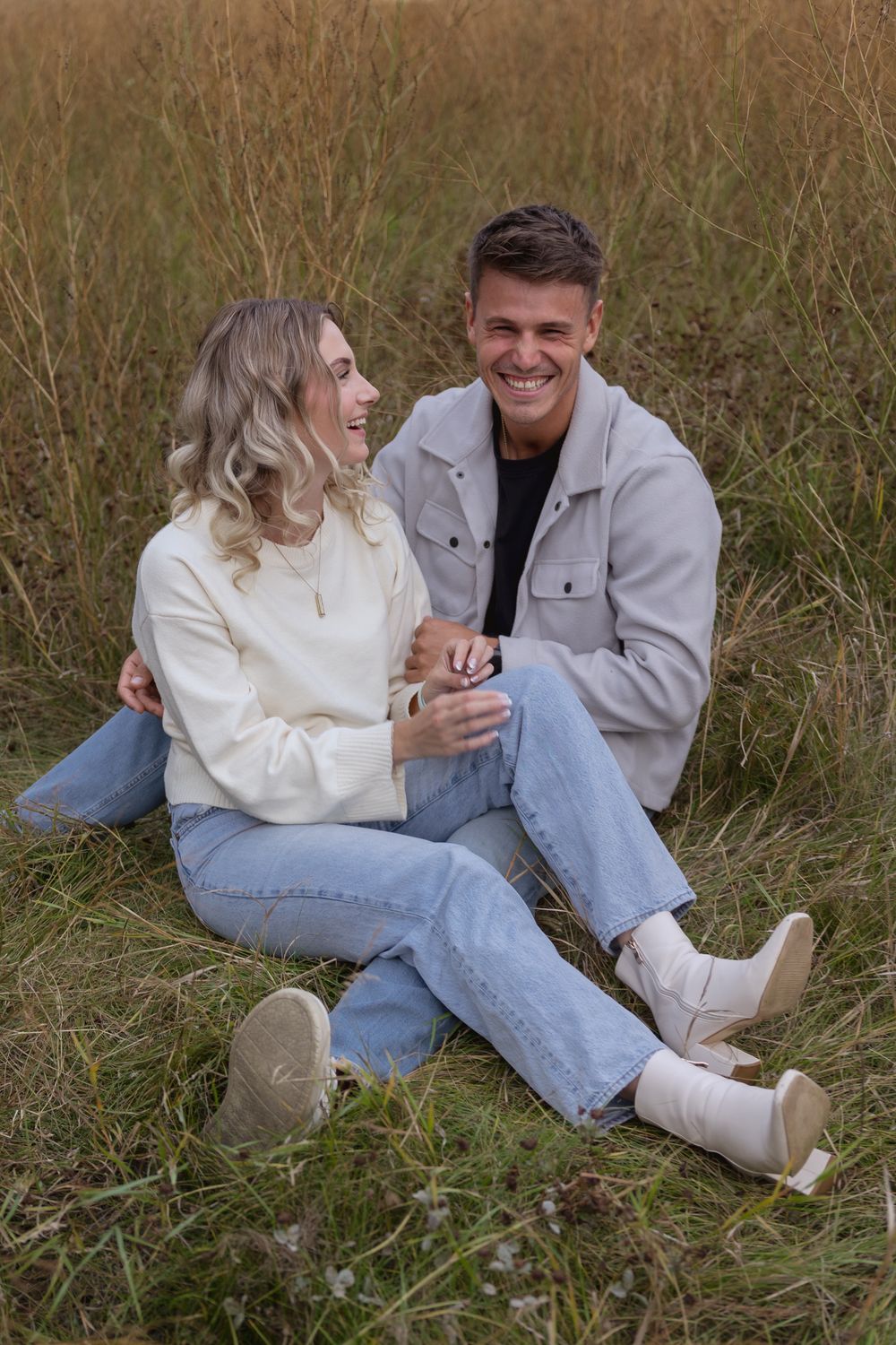 Smiling couple sitting together in a grassy field, wearing casual jeans and light-colored tops, sharing a joyful moment outdoors.