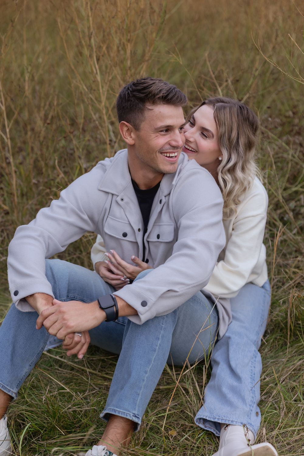 Happy couple sitting in a field, woman whispering to smiling man, both wearing casual denim and light jackets.