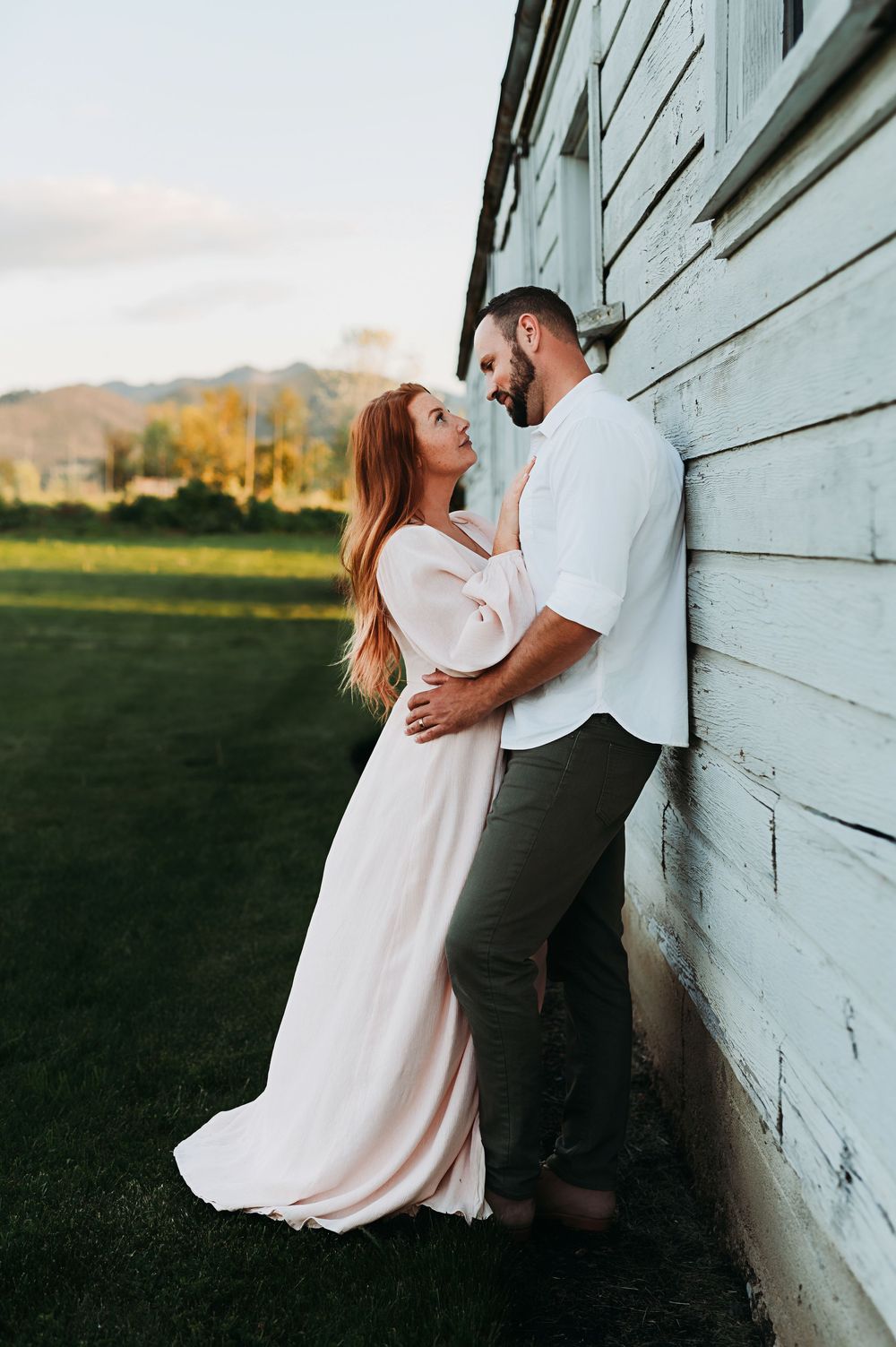 Romantic couple in white and dark clothing embrace against rustic white barn wall at sunset in countryside setting.