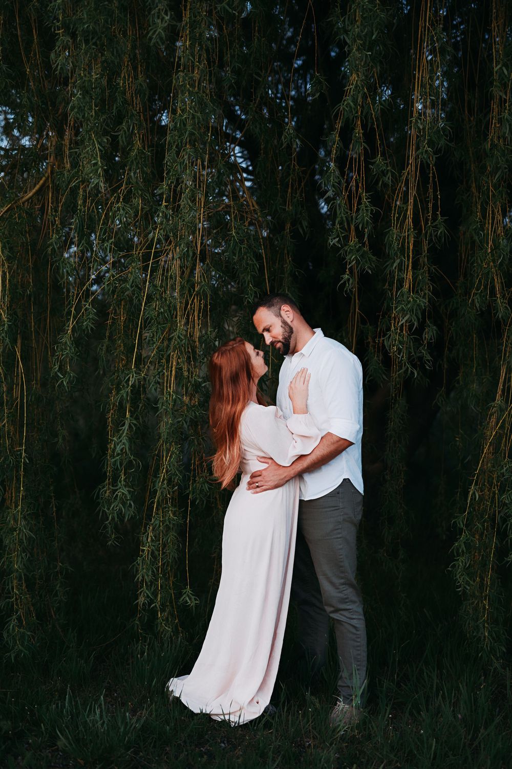 A romantic couple embraces in a wooded area wearing white and grey formal attire during a photo session.