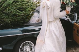 A flowing white winter wedding dress with fur details next to a vintage car.