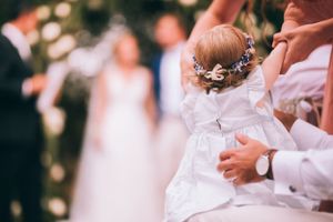Small child in white dress being held during outdoor wedding celebration.
