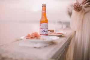 A Budweiser beer bottle sits on a counter alongside a plate in soft pink lighting.