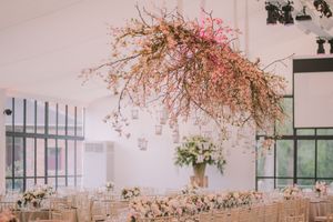 Modern wedding reception venue with suspended floral branch installation and elegant table settings in white and gold decor.