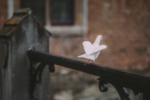 A white paper origami bird perches delicately on a dark metal railing against a blurred brick wall background.