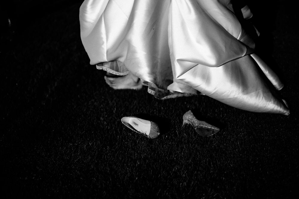 Black and white close-up of wedding dress hem and shoes against dark background.