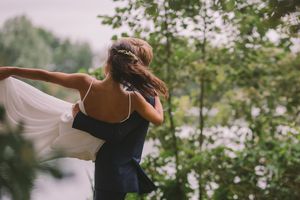 A flowing white dress billows in the breeze as a couple embraces outdoors among green foliage.