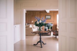 An elegant round table with floral arrangement sits in a bright doorway between classic white interior rooms.